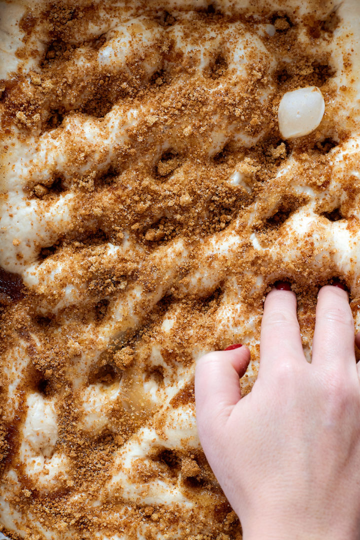A hand presses into a bubbly focaccia dough sprinkled generously with cinnamon sugar, creating dimples for even baking and caramelization.