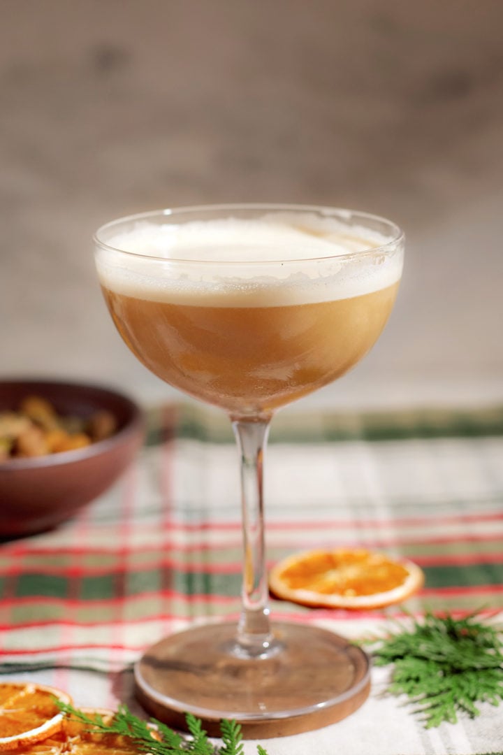 A festive gingerbread whiskey sour cocktail in a coupe glass with a thick foam layer, set on a red and green plaid tablecloth with citrus slices and pine sprigs nearby.