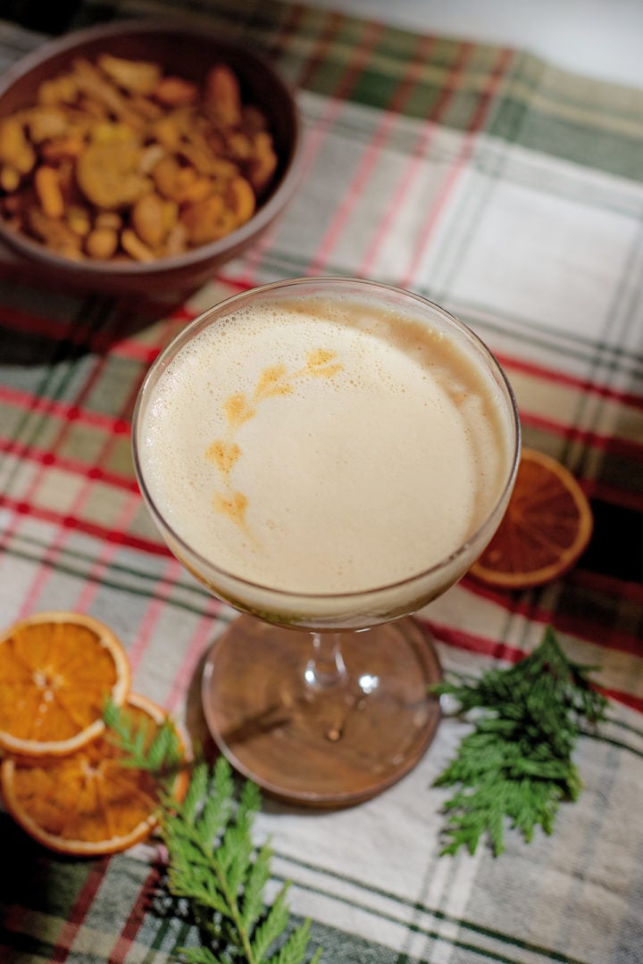 Overhead shot of a gingerbread whiskey sour in a coupe glass, decorated with a heart-shaped aromatic bitters pattern on the foam. Dried oranges and holiday greenery surround the glass.