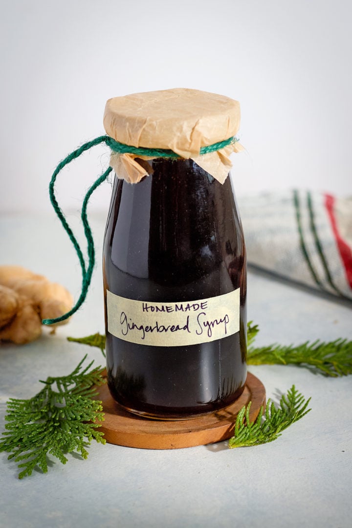 A small glass bottle of homemade gingerbread syrup labeled by hand and wrapped in parchment with green twine. The jar sits on a wooden coaster surrounded by evergreen sprigs and a kitchen towel.