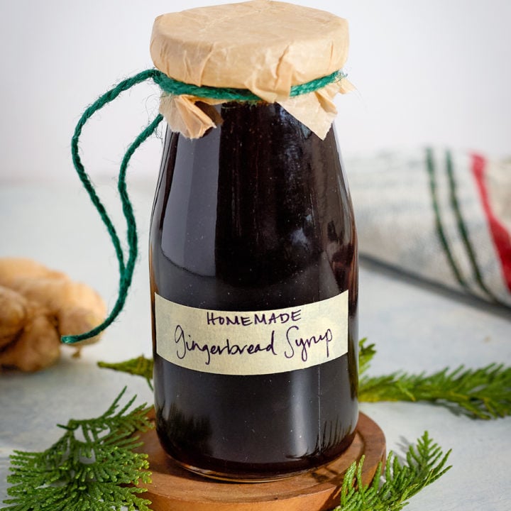 A small glass bottle of homemade gingerbread syrup labeled by hand and wrapped in parchment with green twine. The jar sits on a wooden coaster surrounded by evergreen sprigs and a kitchen towel.