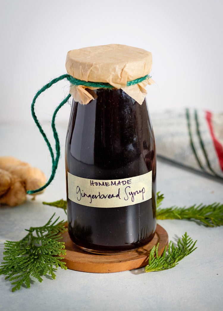 A small glass bottle of homemade gingerbread syrup labeled by hand and wrapped in parchment with green twine. The jar sits on a wooden coaster surrounded by evergreen sprigs and a kitchen towel.