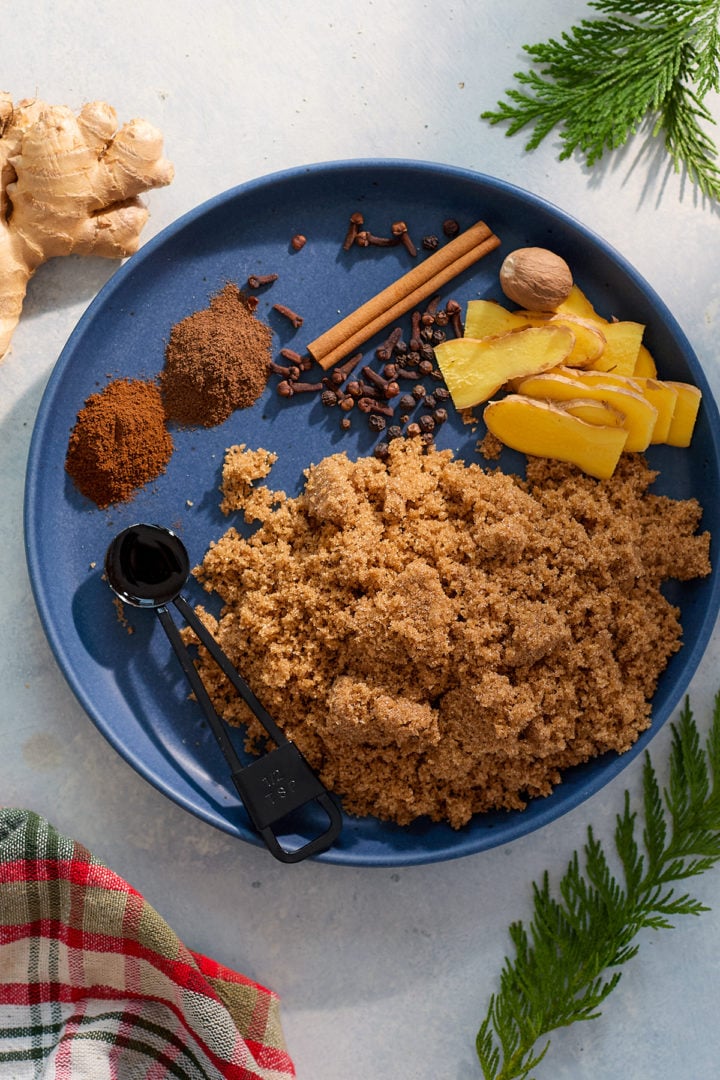 A blue plate filled with gingerbread syrup ingredients: brown sugar, fresh ginger slices, cinnamon sticks, whole cloves, nutmeg, allspice, and a teaspoon of vanilla extract. A festive plaid napkin and pine sprigs are arranged decoratively nearby.