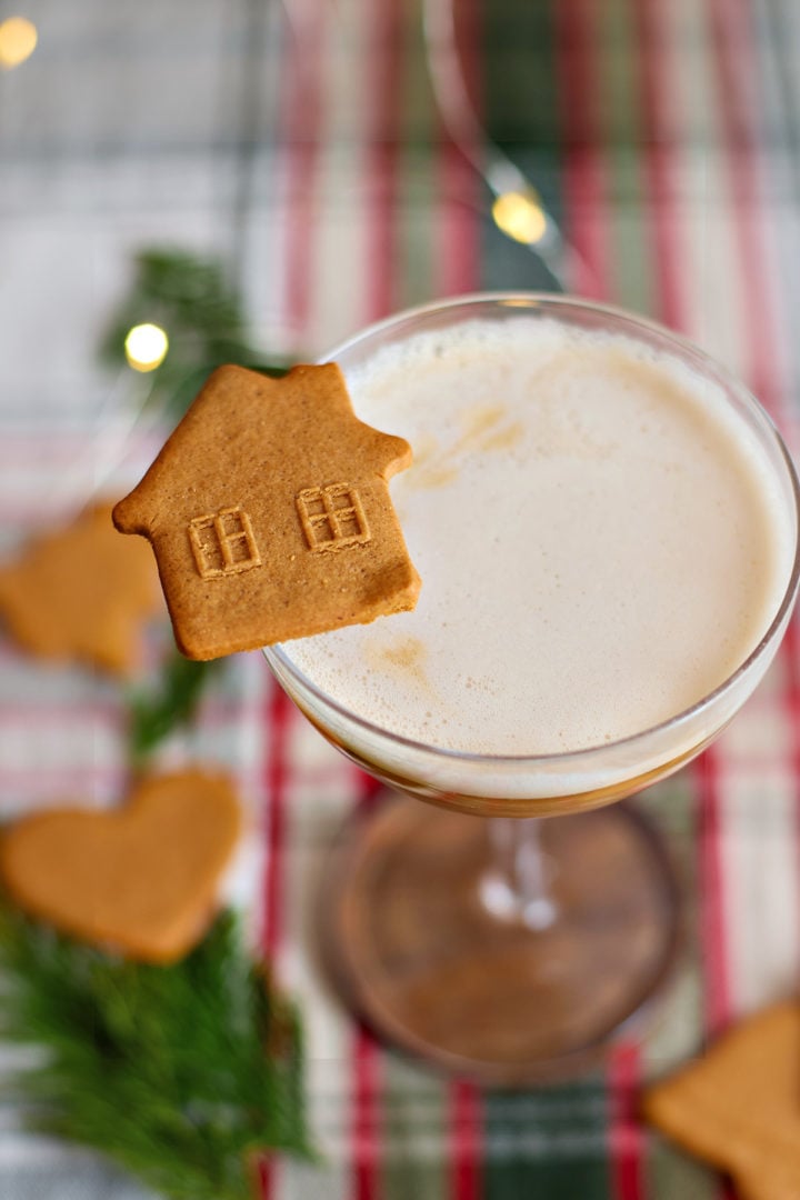 A coupe glass of gingerbread whiskey sour garnished with a house-shaped gingerbread cookie. The background includes heart-shaped cookies, fir sprigs, and warm holiday lights.