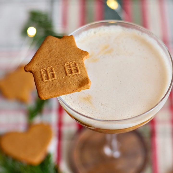 A coupe glass of gingerbread whiskey sour garnished with a house-shaped gingerbread cookie. The background includes heart-shaped cookies, fir sprigs, and warm holiday lights.