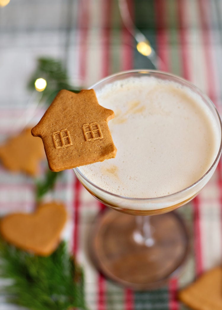 A coupe glass of gingerbread whiskey sour garnished with a house-shaped gingerbread cookie. The background includes heart-shaped cookies, fir sprigs, and warm holiday lights.