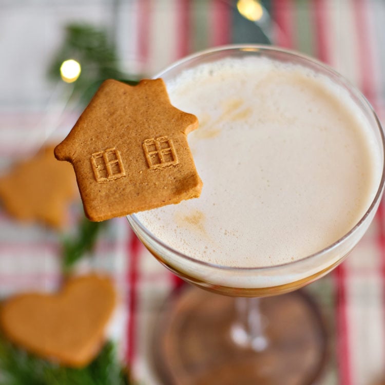 A coupe glass of gingerbread whiskey sour garnished with a house-shaped gingerbread cookie. The background includes heart-shaped cookies, fir sprigs, and warm holiday lights.