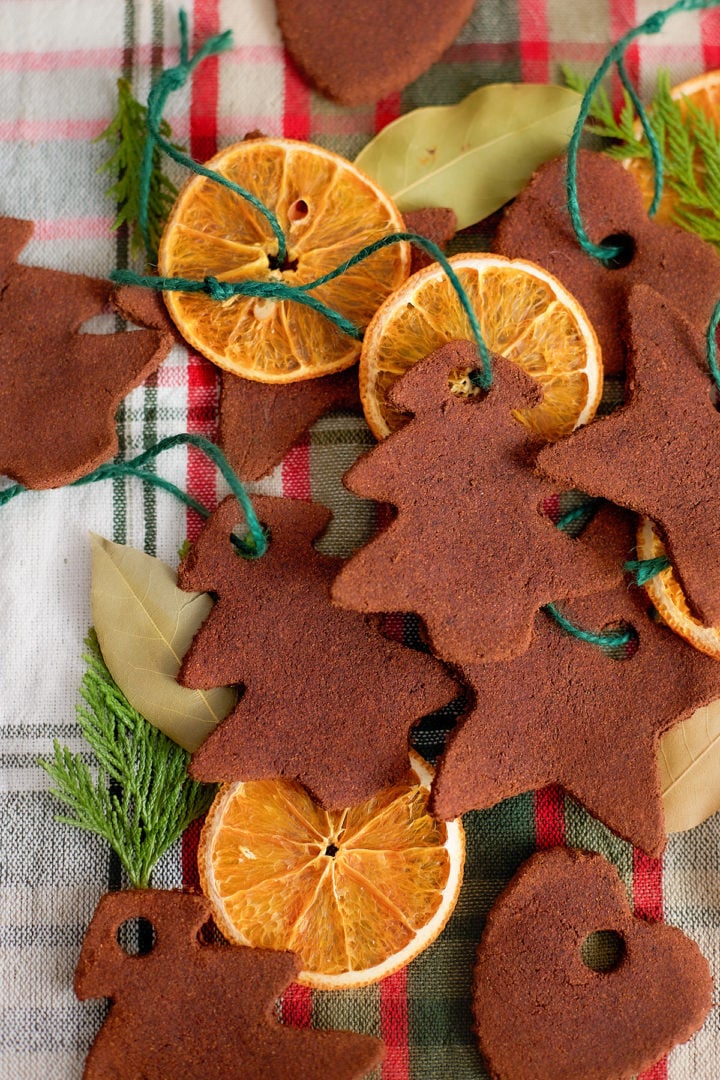 A closer overhead shot of cinnamon dough ornaments in the shapes of trees, stars, and hearts, layered with dried orange slices on a red and green plaid cloth. The ornaments are tied with green string and surrounded by bay leaves and small evergreen clippings.