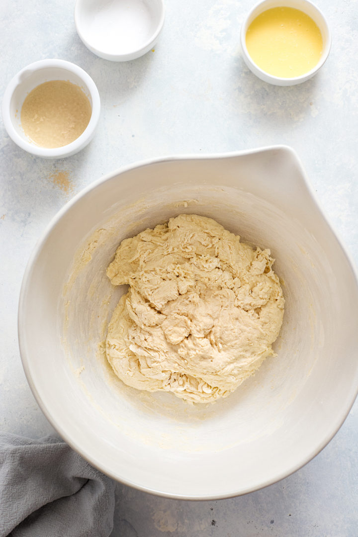 A shaggy mass of freshly mixed dough sits in a large bowl before its first rest, with butter, yeast, and salt nearby, ready for the first rise.