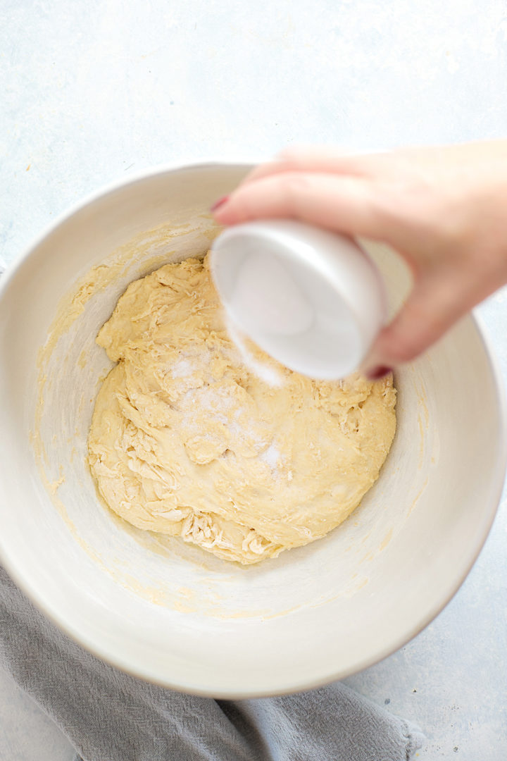 A hand pours a small bowl of fine sea salt over a partially mixed dough in a white mixing bowl during early stages of focaccia preparation.