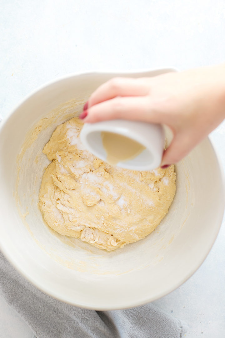 A hand pours dissolved yeast mixture over dough in a white bowl, mid-process during mixing and incorporation of ingredients.
