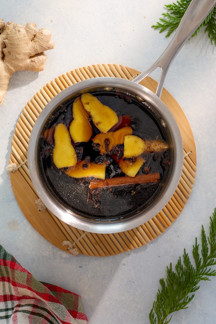 A small saucepan filled with gingerbread syrup in the making, with fresh ginger slices, cinnamon sticks, and spices visible simmering in the dark liquid. The pot rests on a bamboo trivet with festive kitchen decor nearby.
