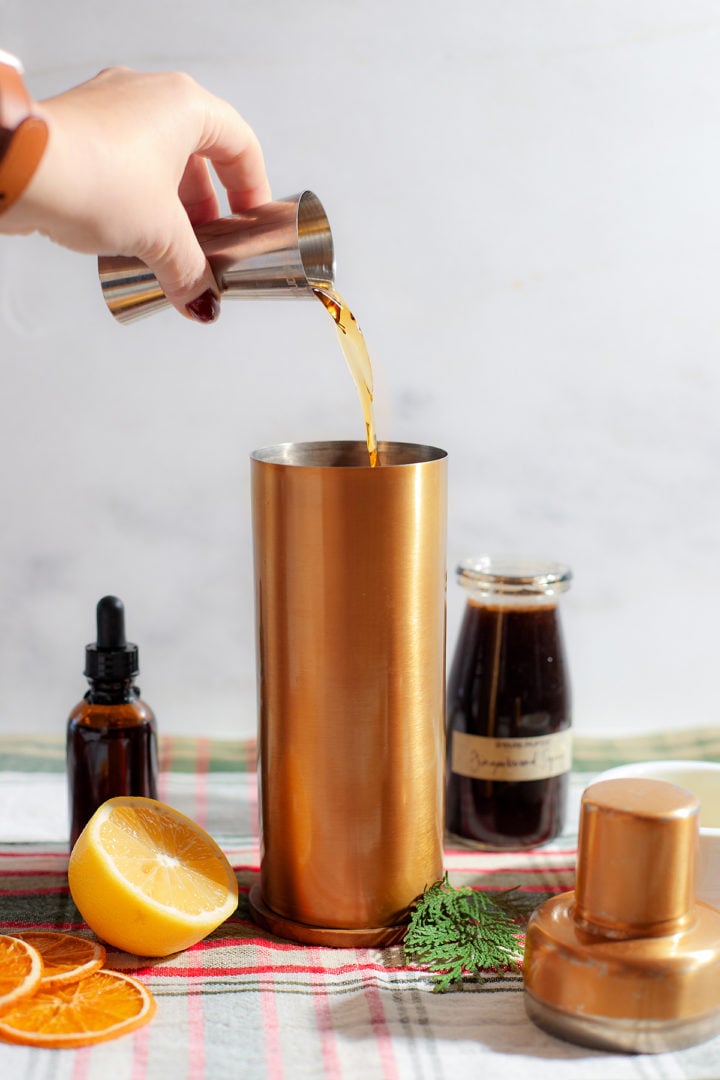 A hand pouring whiskey from a jigger into a gold cocktail shaker. Ingredients including a halved lemon, gingerbread syrup, and a bitters bottle are arranged around it.