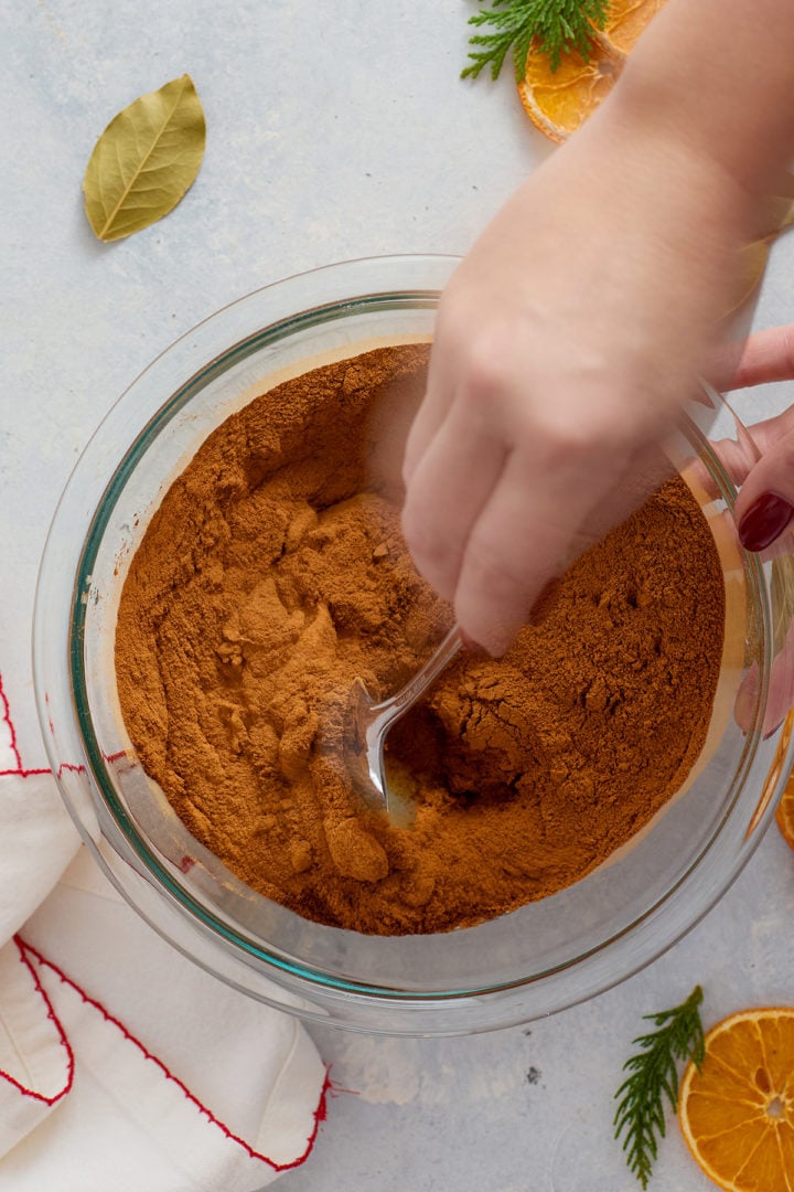 A person’s hand is using a metal spoon to stir a large amount of ground cinnamon into a mixing bowl containing wet ingredients. Only the hand, spoon, and part of the cinnamon mixture are visible. Bay leaves, dried oranges, and greenery decorate the surrounding surface.