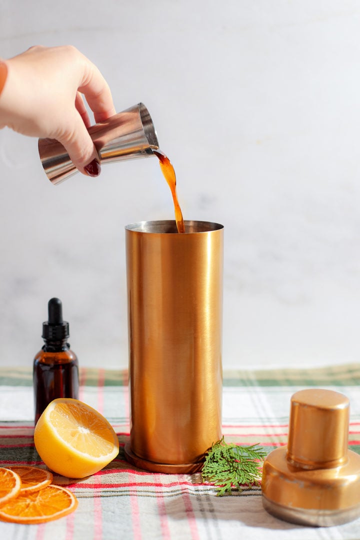 Gingerbread simple syrup being poured from a jigger into a cocktail shaker, with holiday-themed ingredients and décor surrounding the shaker.