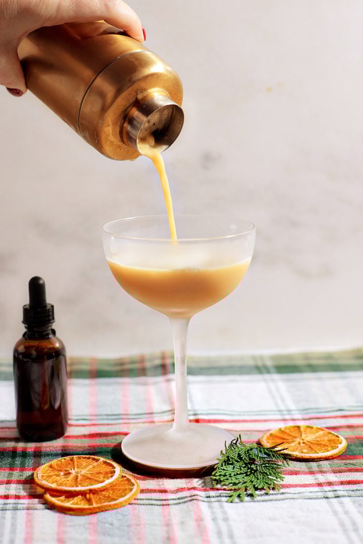 A hand pouring the frothy gingerbread whiskey sour from a golden shaker into a coupe glass. Garnishes include dried orange slices and pine sprigs on a festive plaid cloth.