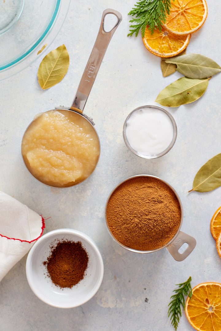 Overhead photo of a flat lay arrangement showing ingredients in metal and ceramic measuring cups: one cup of applesauce, one cup of white liquid glue, one cup of ground cinnamon, and a small bowl of ground cloves. The ingredients are placed on a light surface with scattered dried orange slices, bay leaves, and evergreen sprigs.