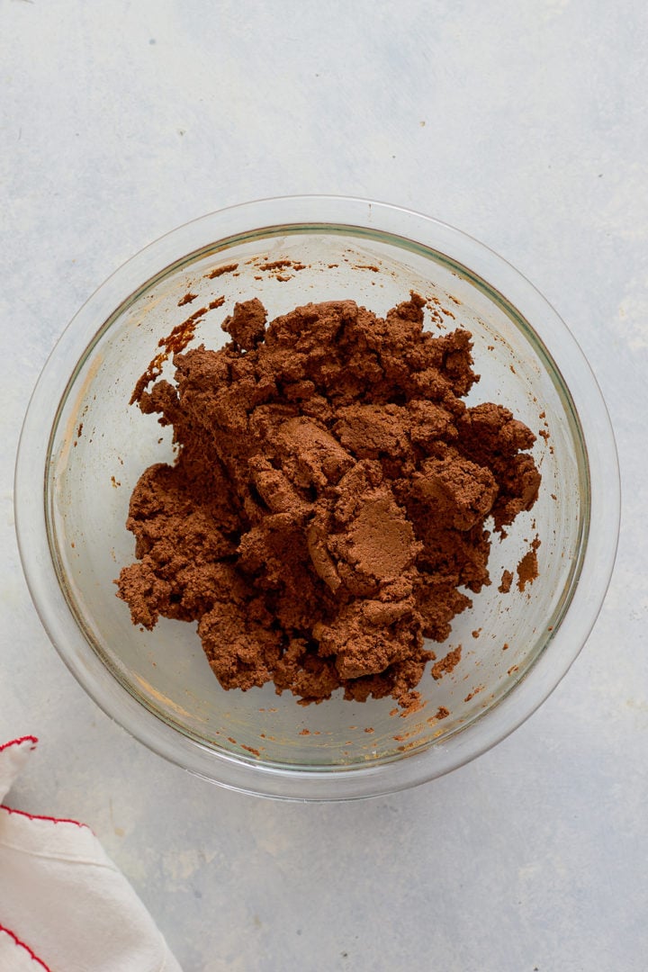 A glass mixing bowl filled with brown cinnamon dough sits on a light surface. The dough appears thick and slightly crumbly, sticking to the sides of the bowl. A white cloth with red stitching is partially visible in the bottom corner.