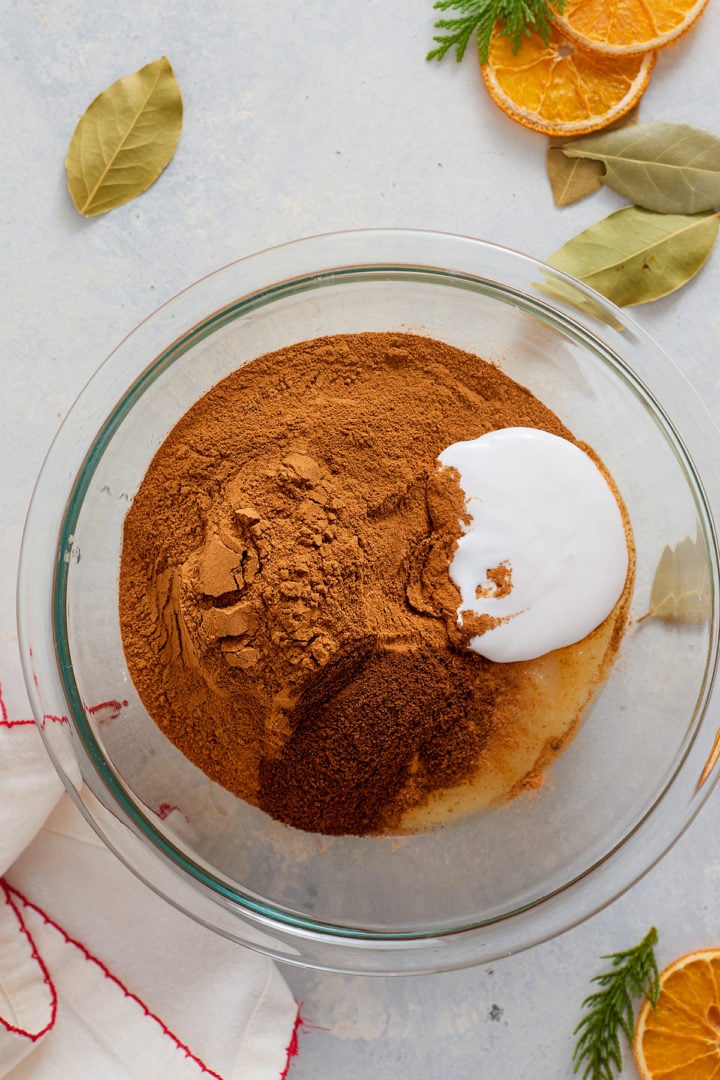 A clear glass mixing bowl contains a mound of unmixed ingredients: ground cinnamon, white liquid glue, and applesauce. The mixture is not yet stirred. Bay leaves, dried orange slices, and evergreen clippings are scattered on the light-colored surface around the bowl.